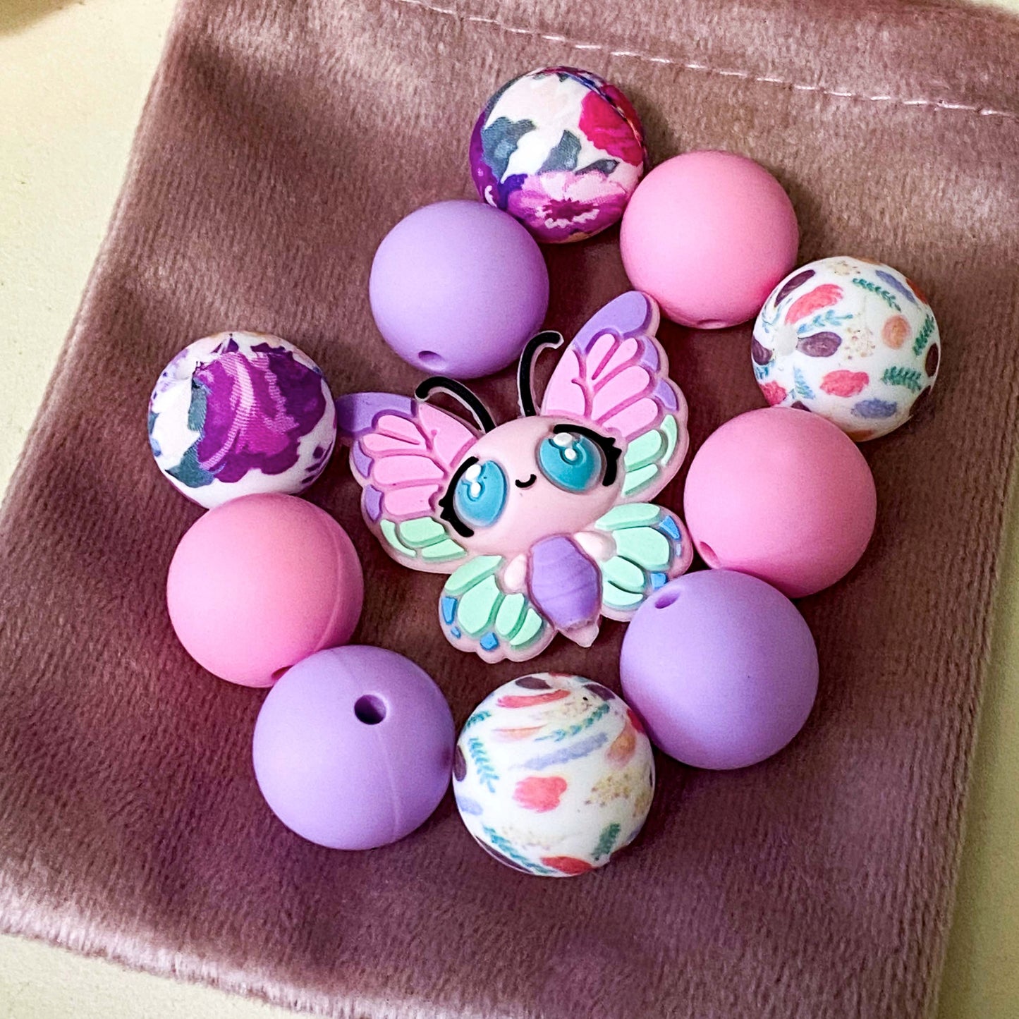 pastel silicone bead bundle with butterfly feature bead, pink and lilac round beads and floral patterned beads on a velvet bag