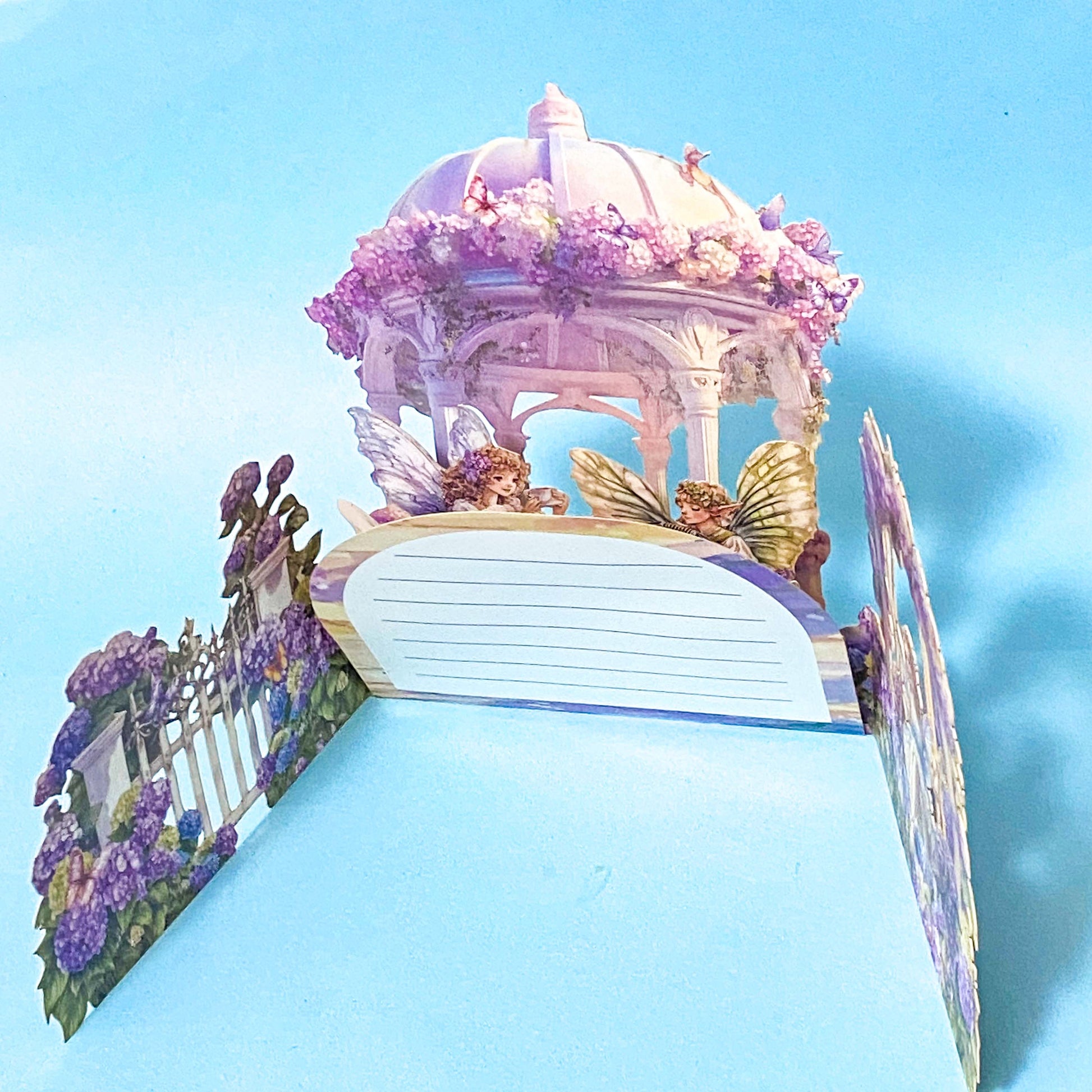 Decorative pop up 3d card with a fairy garden design featuring a gazebo, butterflies, and flowers on a blue background.