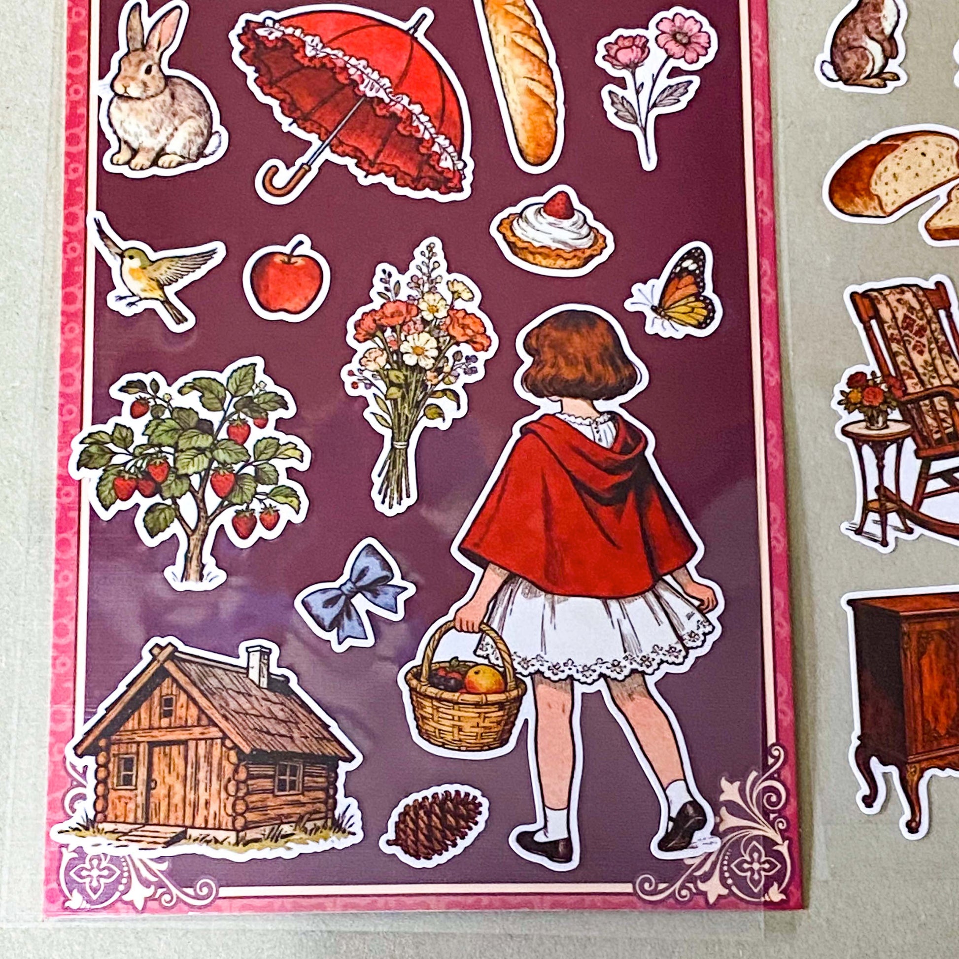 red riding hood's cottage matte sticker pack with two decorative clear sheets of stickers featuring little red riding hood, woodland animals, cottages, flowers, baskets, books, strawberries, squirrels, rabbits and cosy cottagecore illustrations.