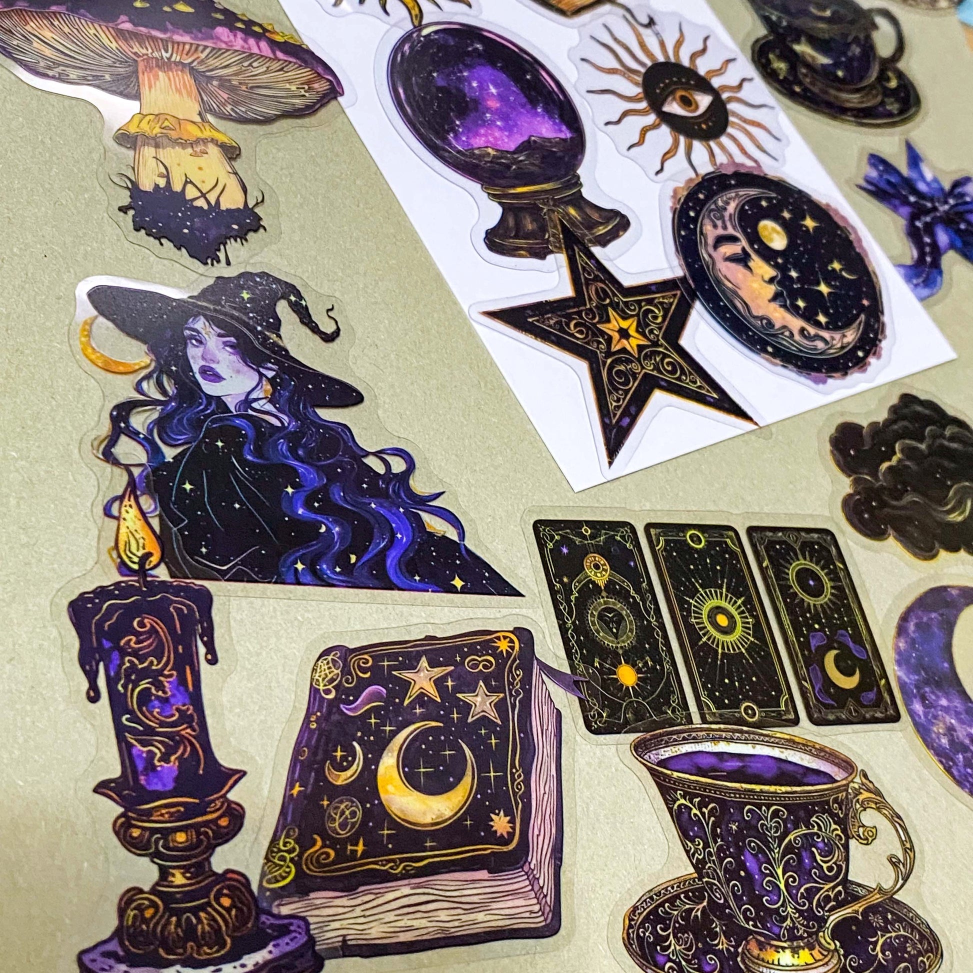 pack of 50 transparent PET witchy stickers featuring celestial sun and moon, crystal balls, spell books, ornate teacups, witches, black cats, magical eyes and vintage style mystical illustrations in deep blue, purple, black and gold. 