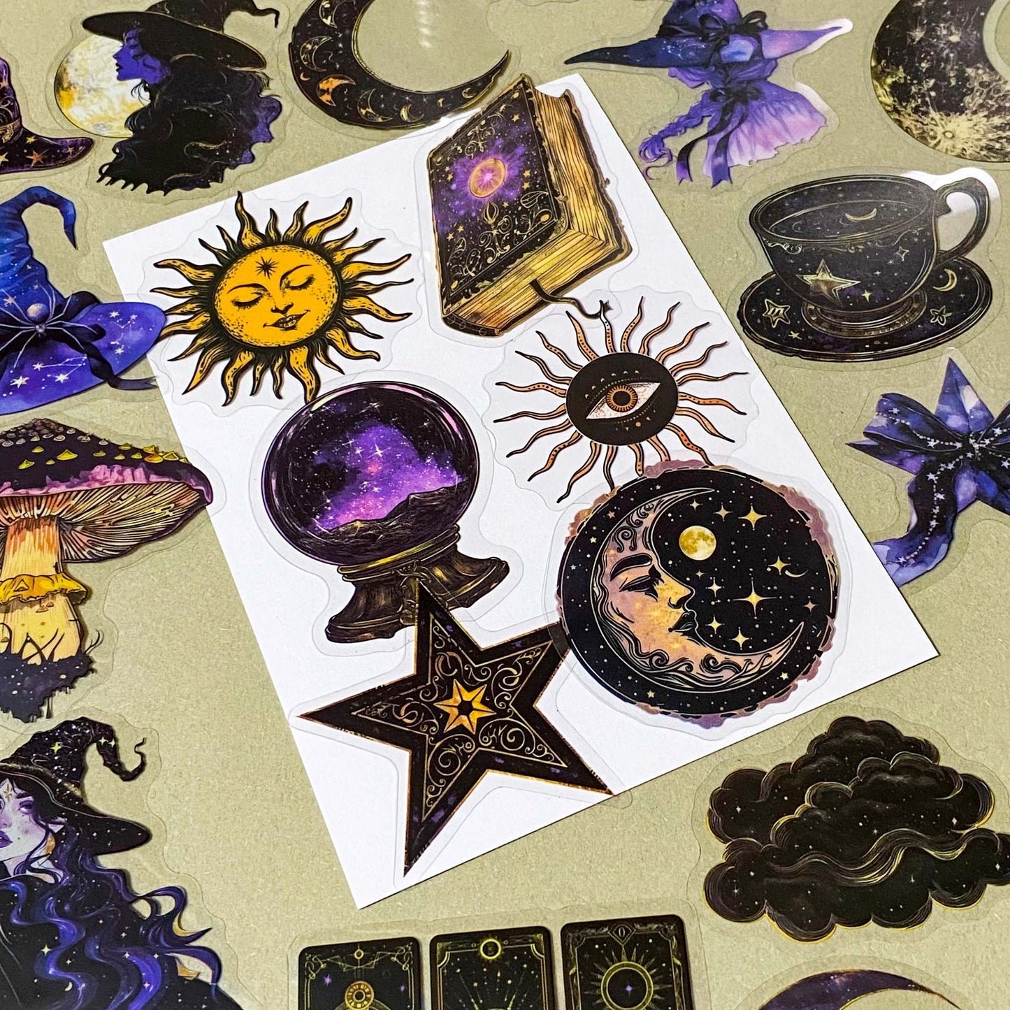 pack of 50 transparent PET witchy stickers featuring celestial sun and moon, crystal balls, spell books, ornate teacups, witches, black cats, magical eyes and vintage style mystical illustrations in deep blue, purple, black and gold. 