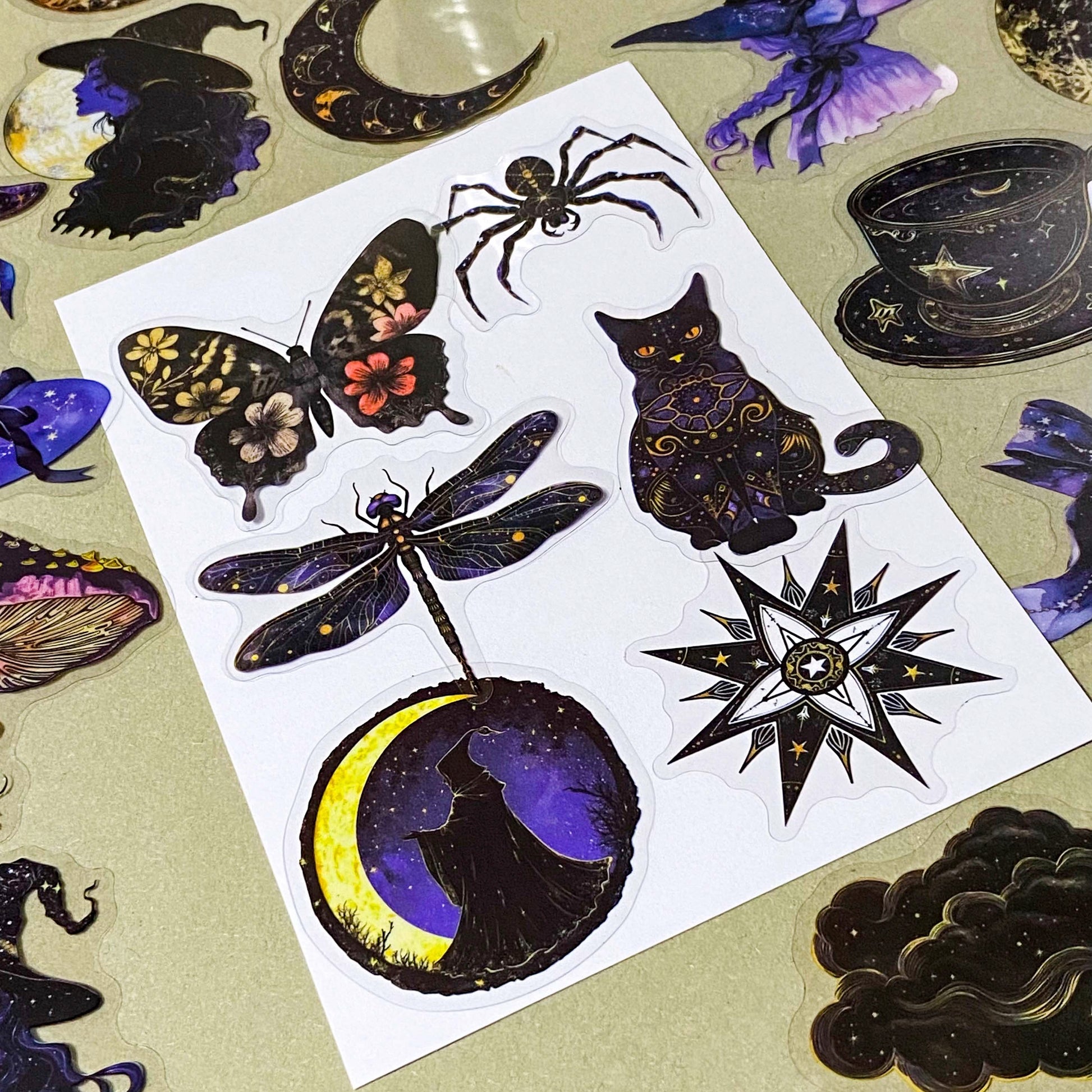 pack of 50 transparent PET witchy stickers featuring celestial sun and moon, crystal balls, spell books, ornate teacups, witches, black cats, magical eyes and vintage style mystical illustrations in deep blue, purple, black and gold. 