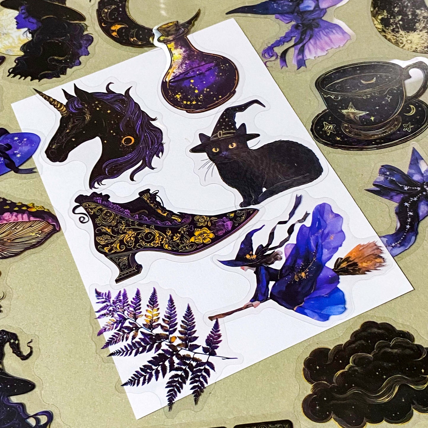 pack of 50 transparent PET witchy stickers featuring celestial sun and moon, crystal balls, spell books, ornate teacups, witches, black cats, magical eyes and vintage style mystical illustrations in deep blue, purple, black and gold. 