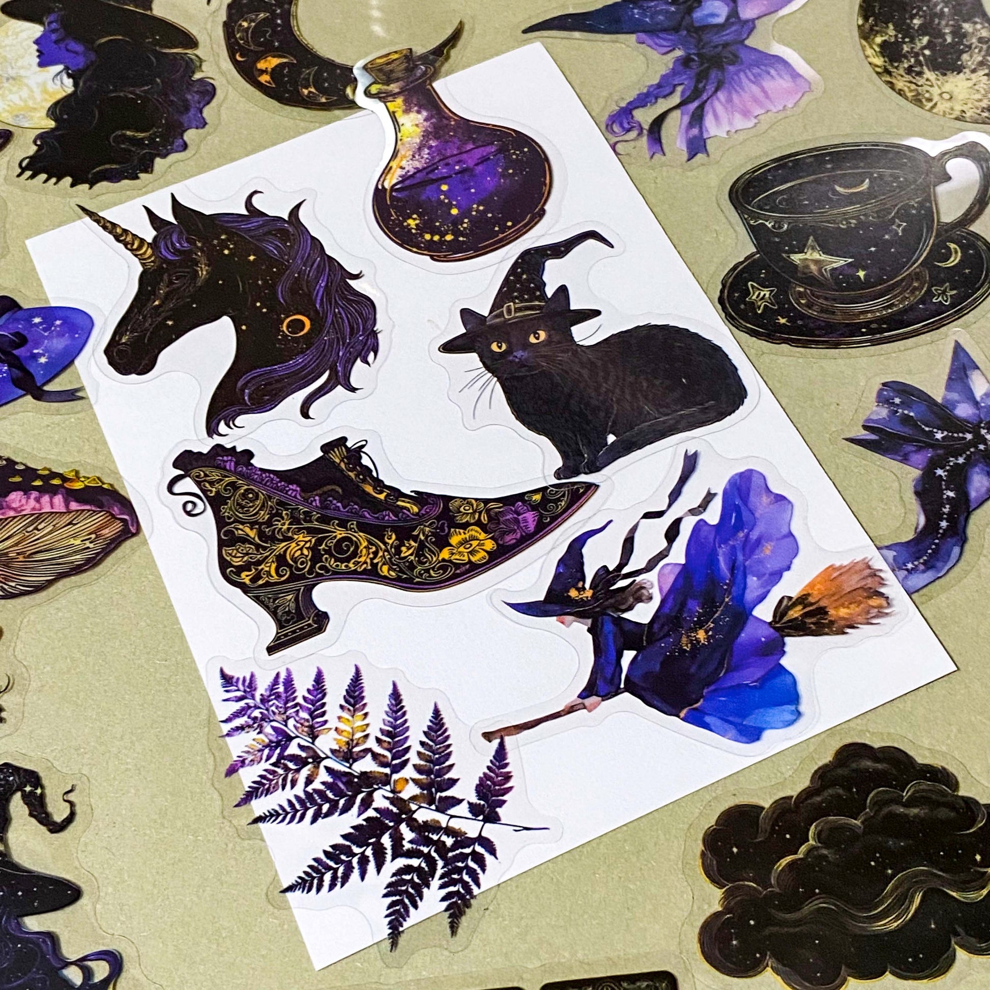 pack of 50 transparent PET witchy stickers featuring celestial sun and moon, crystal balls, spell books, ornate teacups, witches, black cats, magical eyes and vintage style mystical illustrations in deep blue, purple, black and gold. 