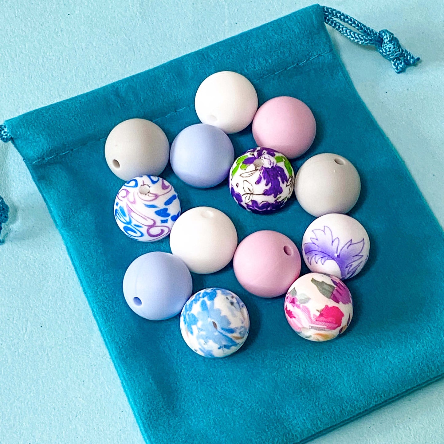 silicone 16mm beads bundle with floral patterned beads and coordinating plain beads in pale pink, light blue, grey and white on a teal velvet bag