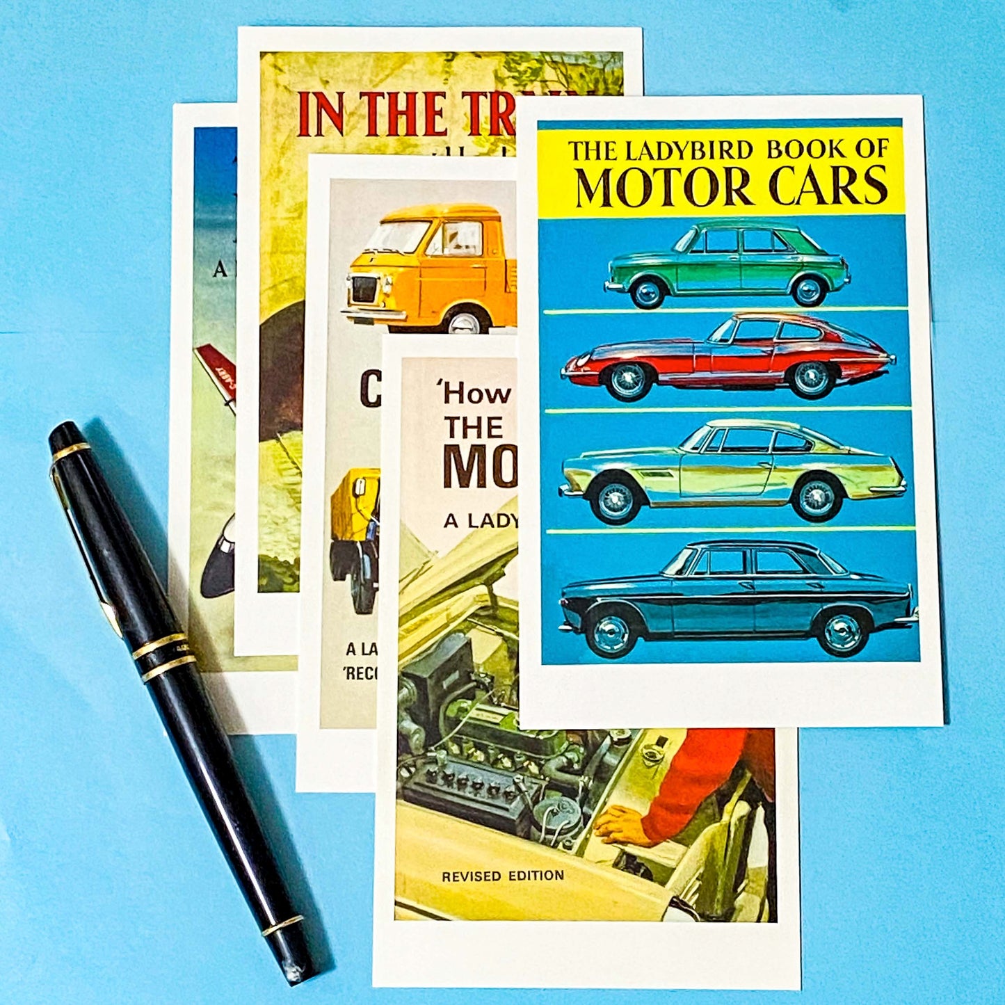 Collection of Ladybird book covers postcards on motor cars and transport with a pen on a blue background