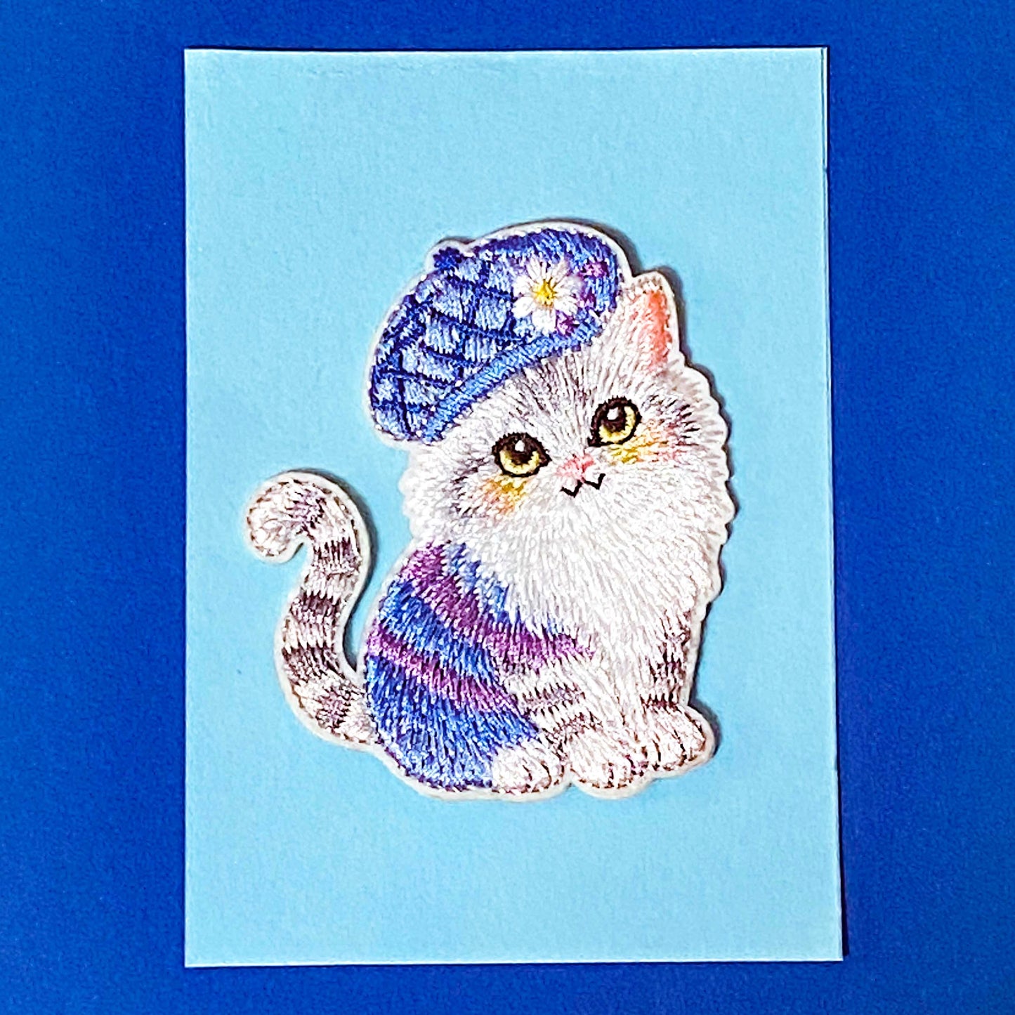 Embroidered fabric patch of a cat wearing a colorful hat on a blue card