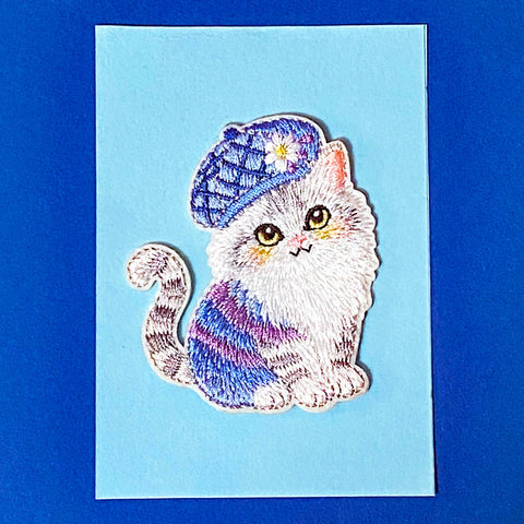 Embroidered fabric patch of a cat wearing a colorful hat on a blue card