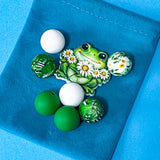 green frog bead bundle with double sided jumbo daisy frog ceramic focal bead and coordinating floral, white and deep green silicone beads on bright blue background