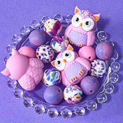 pastel owl silicone bead bundle with marbled purple beads and floral pink beads arranged on a glass plate on a purple background