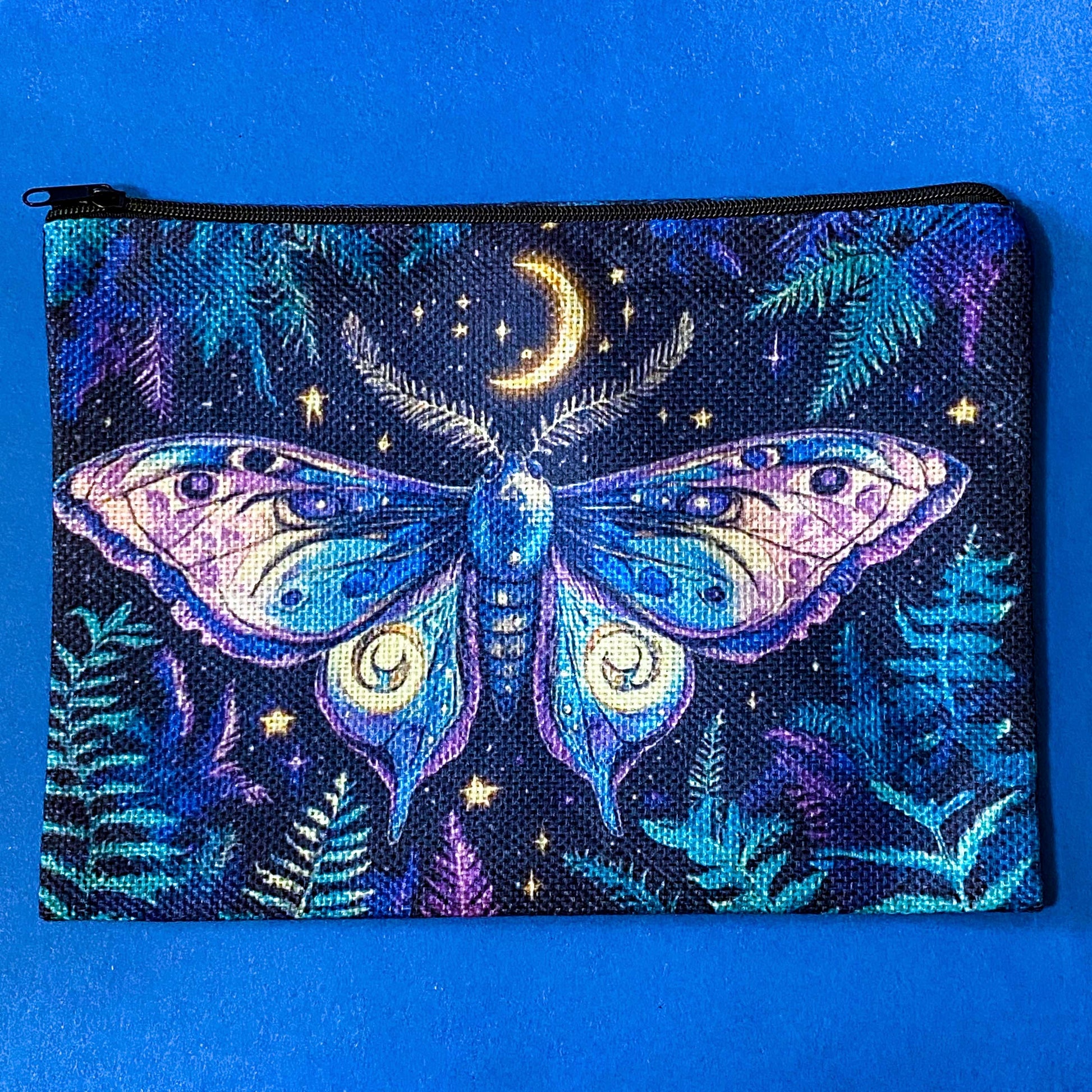 A zippered pouch featuring a detailed fantasy print of a large moth under a crescent moon and stars, set against a dark background with fern motifs, displayed on a solid blue background.