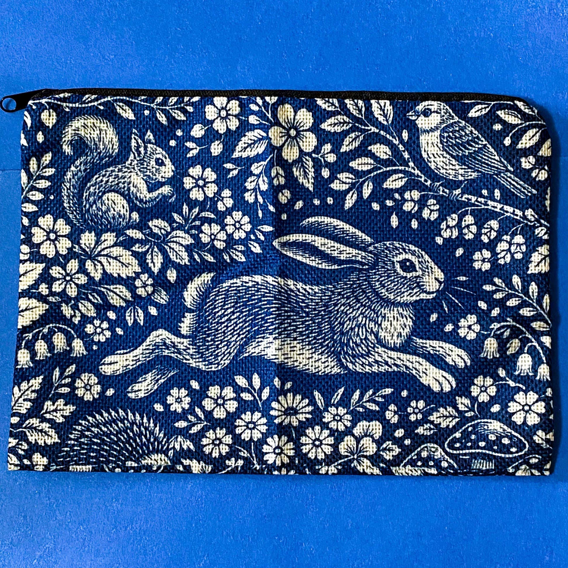 navy blue zip pouch made of canvas with a deep blue and cream design featuring a running hare, squirrel, bird and hedgehog among flowers and mushrooms in a forest scene. Uk letterbox gifts.