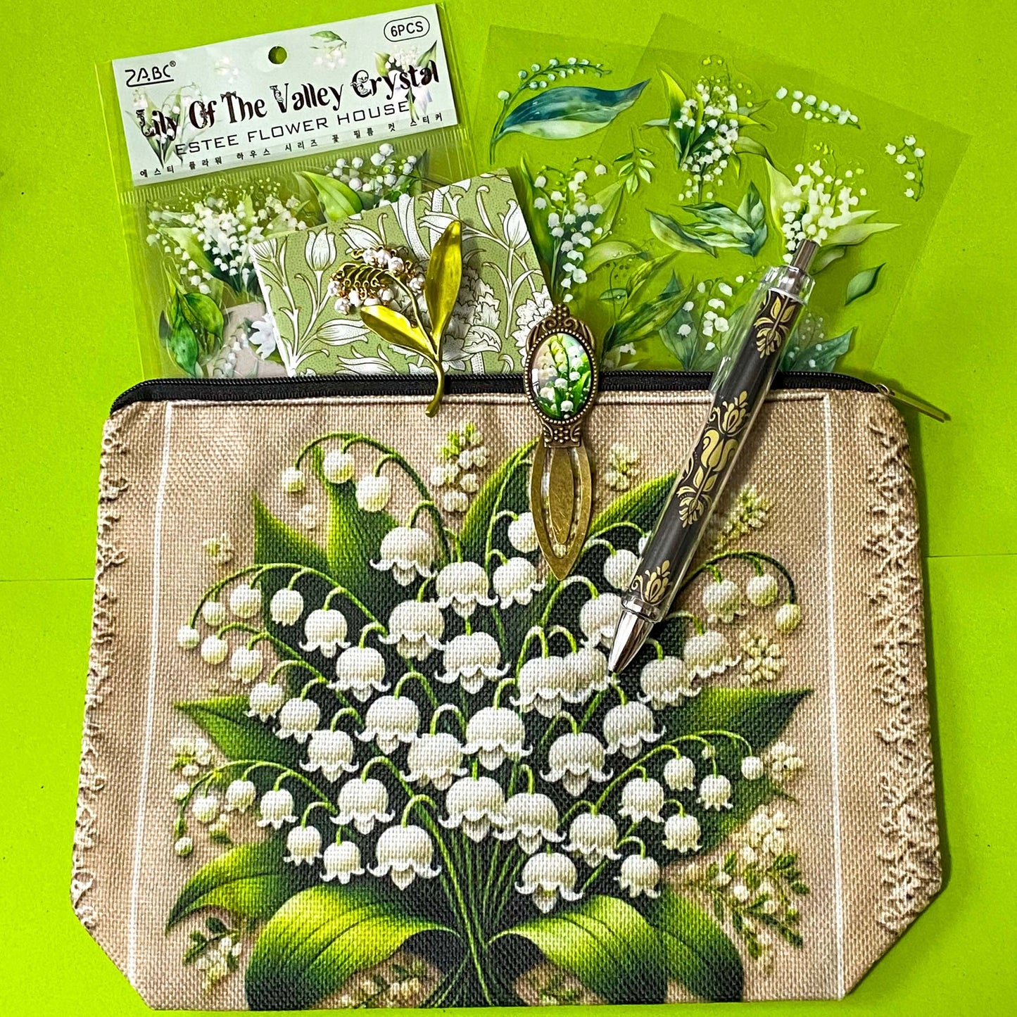 lily of the valley botanical gift set including floral zip canvas pouch, articulated lily of the valley metal brooch, botanical glass cabochon bookmark, floral mini notebook, black ornate ballpoint pen and pack of 6 clear PET lily of the valley large sticker sheets. Green, natural cream and white coloured set. Uk letterbox gift shop for vintage and beautiful gifts and keepsakes.