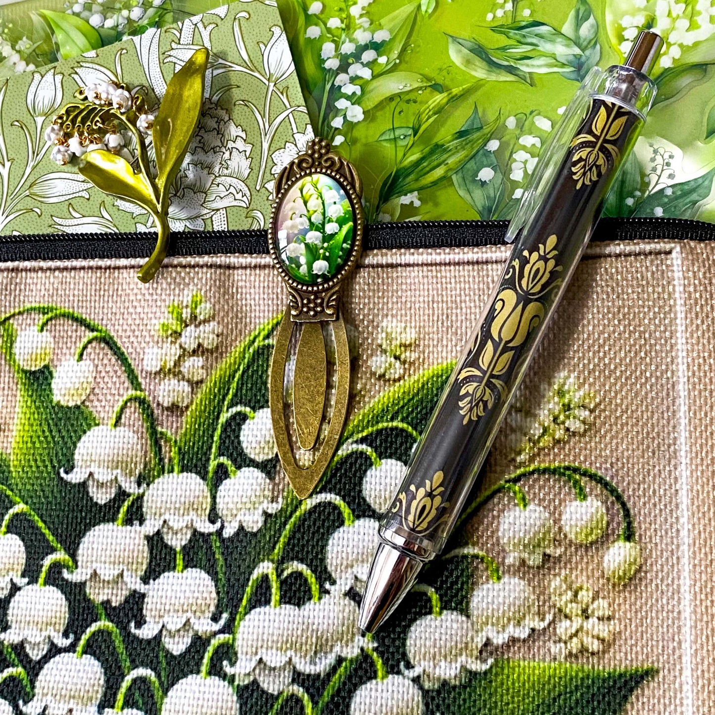 lily of the valley botanical gift set including floral zip canvas pouch, articulated lily of the valley metal brooch, botanical glass cabochon bookmark, floral mini notebook, black ornate ballpoint pen and pack of 6 clear PET lily of the valley large sticker sheets. Green, natural cream and white coloured set. Uk letterbox gift shop for vintage and beautiful gifts and keepsakes.