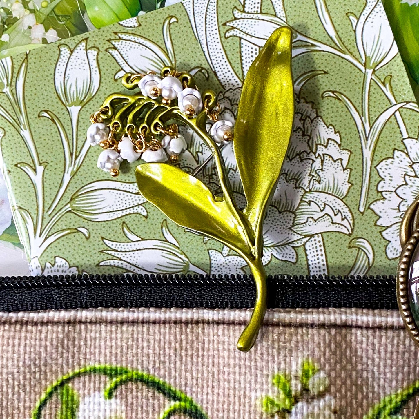 lily of the valley botanical gift set including floral zip canvas pouch, articulated lily of the valley metal brooch, botanical glass cabochon bookmark, floral mini notebook, black ornate ballpoint pen and pack of 6 clear PET lily of the valley large sticker sheets. Green, natural cream and white coloured set. Uk letterbox gift shop for vintage and beautiful gifts and keepsakes.