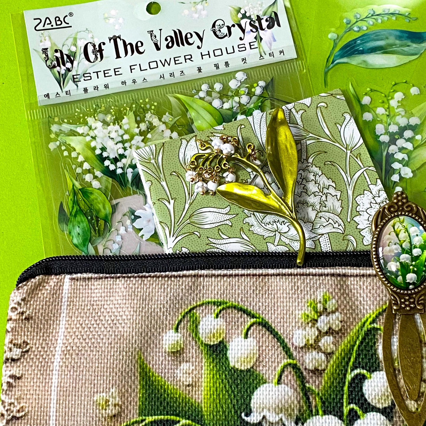 lily of the valley botanical gift set including floral zip canvas pouch, articulated lily of the valley metal brooch, botanical glass cabochon bookmark, floral mini notebook, black ornate ballpoint pen and pack of 6 clear PET lily of the valley large sticker sheets. Green, natural cream and white coloured set. Uk letterbox gift shop for vintage and beautiful gifts and keepsakes.