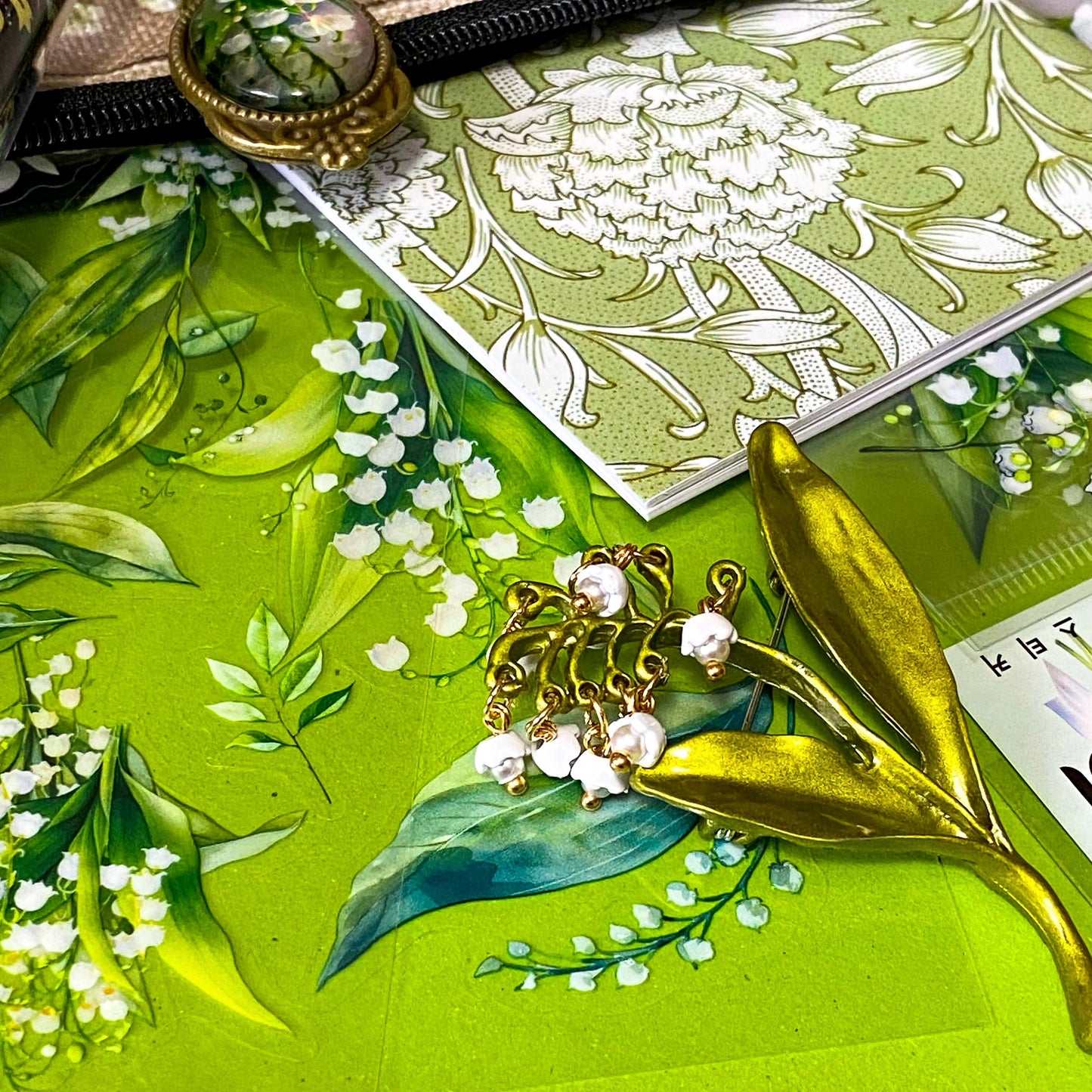 lily of the valley botanical gift set including floral zip canvas pouch, articulated lily of the valley metal brooch, botanical glass cabochon bookmark, floral mini notebook, black ornate ballpoint pen and pack of 6 clear PET lily of the valley large sticker sheets. Green, natural cream and white coloured set. Uk letterbox gift shop for vintage and beautiful gifts and keepsakes.