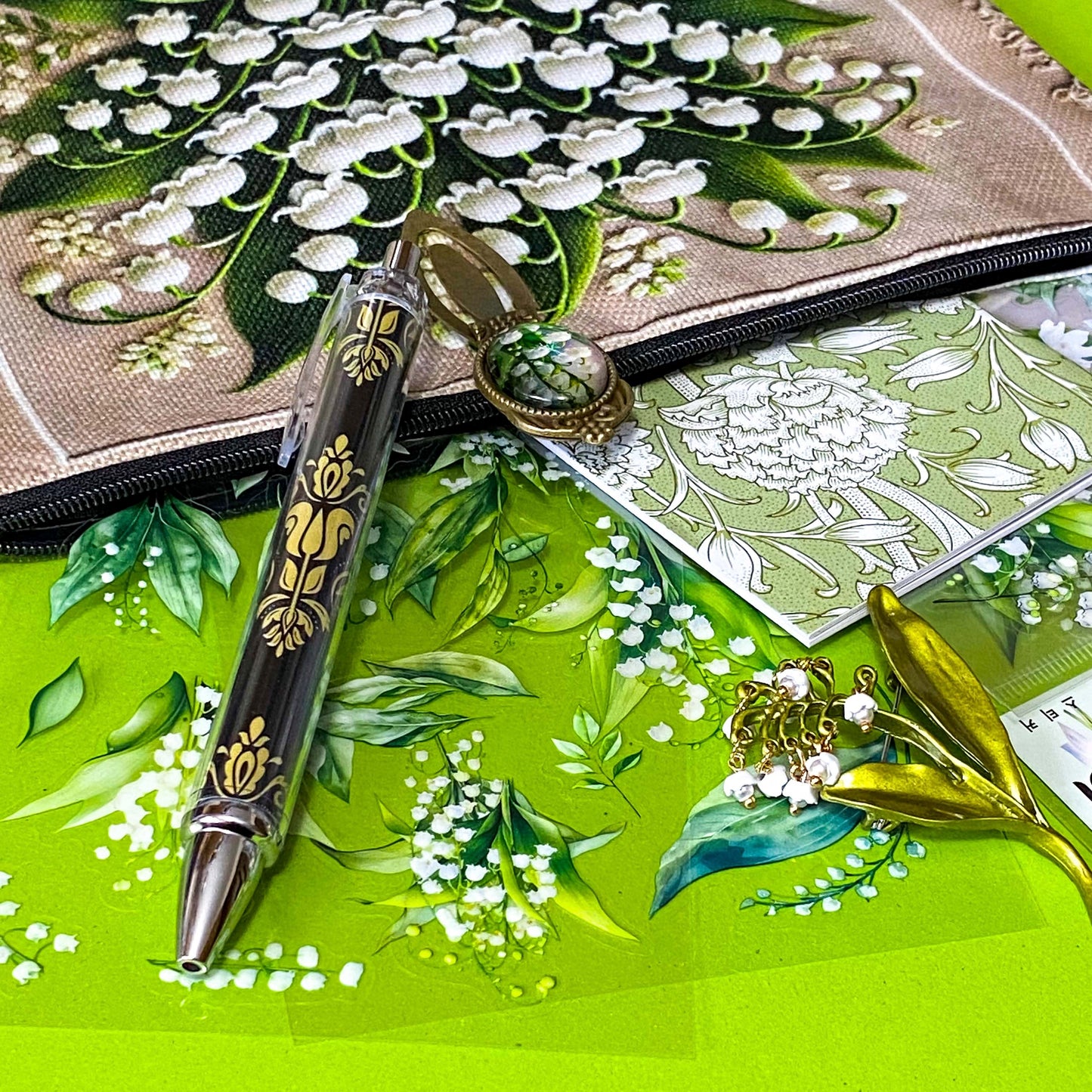 lily of the valley botanical gift set including floral zip canvas pouch, articulated lily of the valley metal brooch, botanical glass cabochon bookmark, floral mini notebook, black ornate ballpoint pen and pack of 6 clear PET lily of the valley large sticker sheets. Green, natural cream and white coloured set. Uk letterbox gift shop for vintage and beautiful gifts and keepsakes.