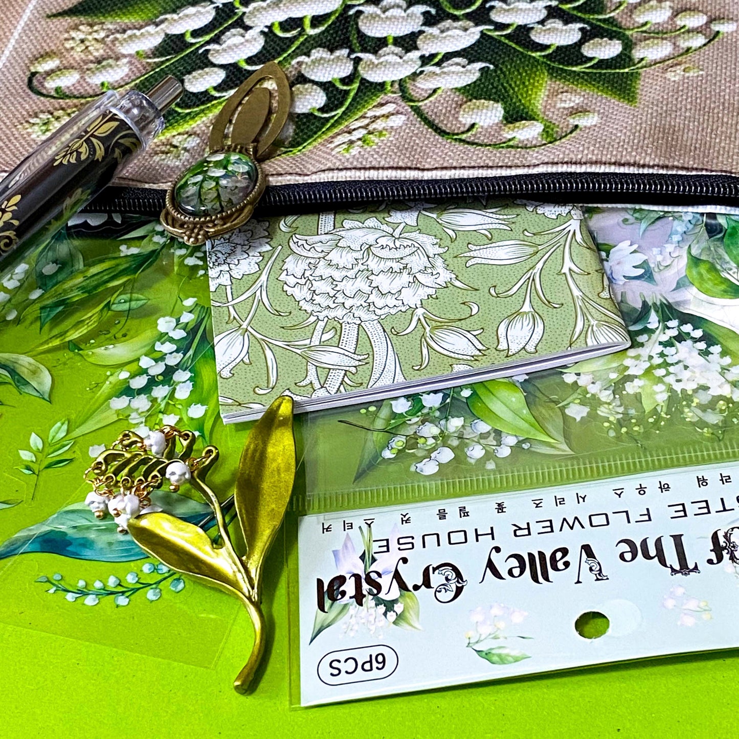 lily of the valley botanical gift set including floral zip canvas pouch, articulated lily of the valley metal brooch, botanical glass cabochon bookmark, floral mini notebook, black ornate ballpoint pen and pack of 6 clear PET lily of the valley large sticker sheets. Green, natural cream and white coloured set. Uk letterbox gift shop for vintage and beautiful gifts and keepsakes.