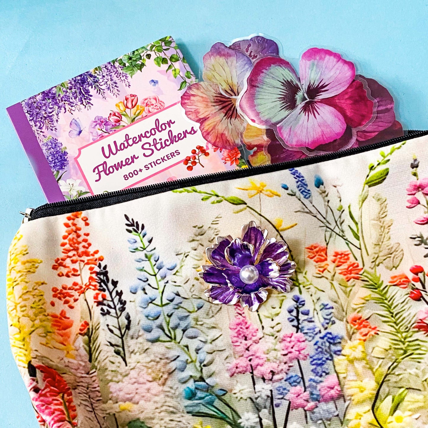 floral gift set with illustrated flower pouch, watercolour sticker book and purple enamel flower brooch on a blue background