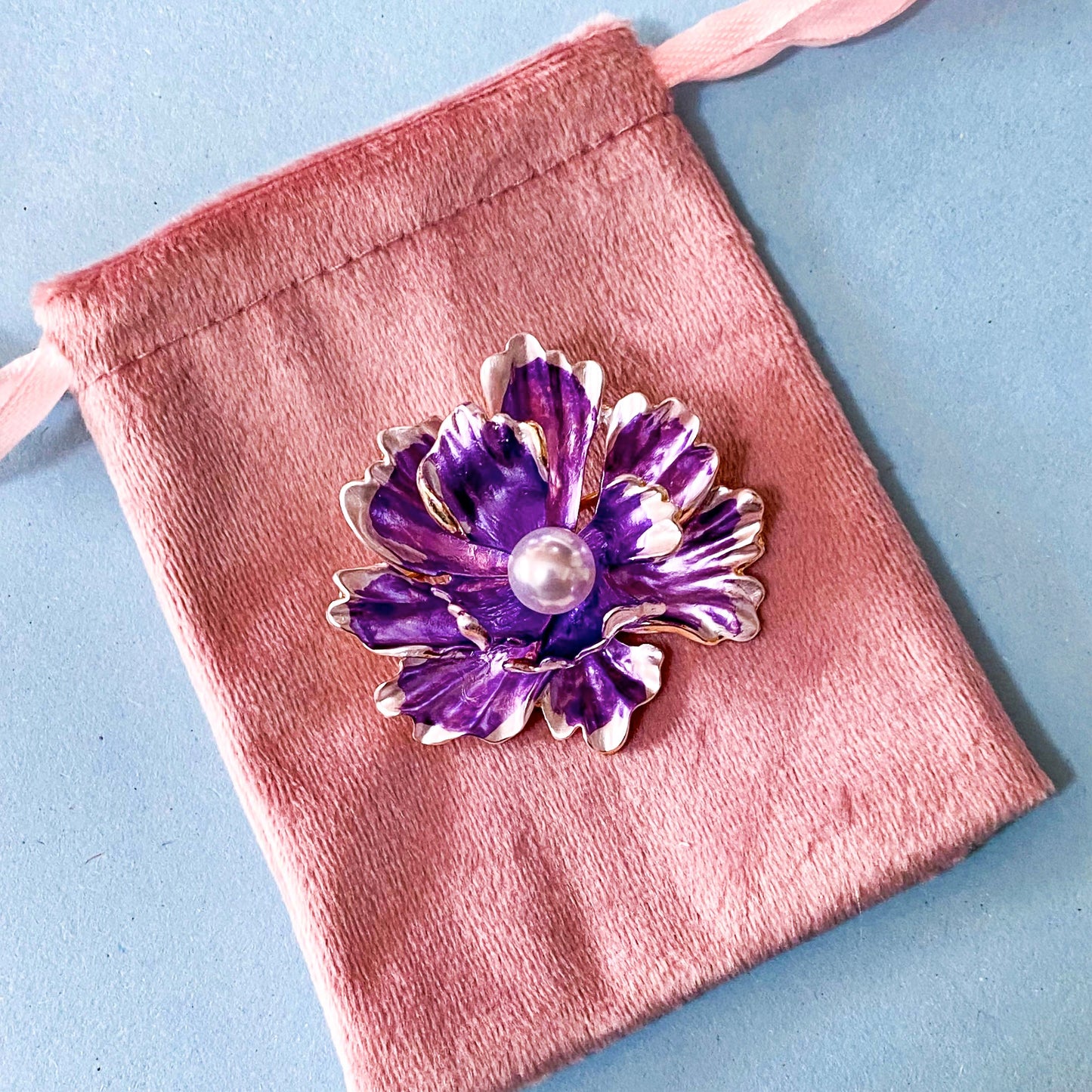 Purple flower-shaped brooch with pearl center on a pink velvet pouch against a blue background
