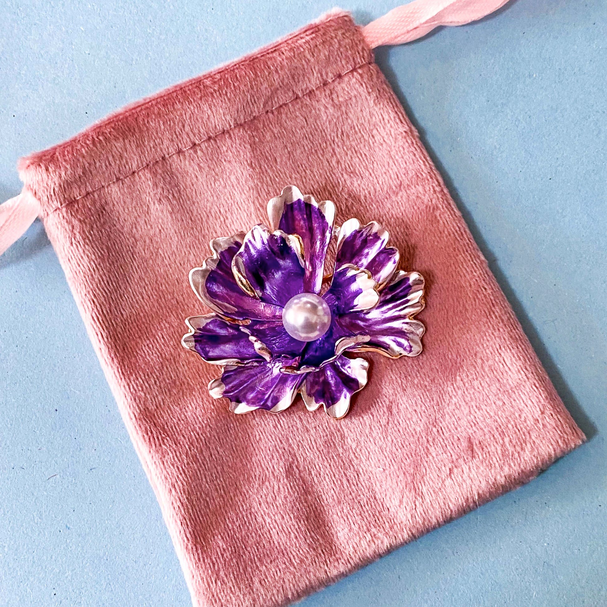 Purple flower-shaped brooch with pearl center on a pink velvet pouch against a blue background