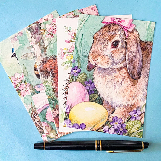 set of 6 spring animal postcards featuring bunny, blue bird, bee, squirrel and hedgehog illustrations with blossom flowers, wrapped in a kraft belly band with wax seal sticker on a soft blue background