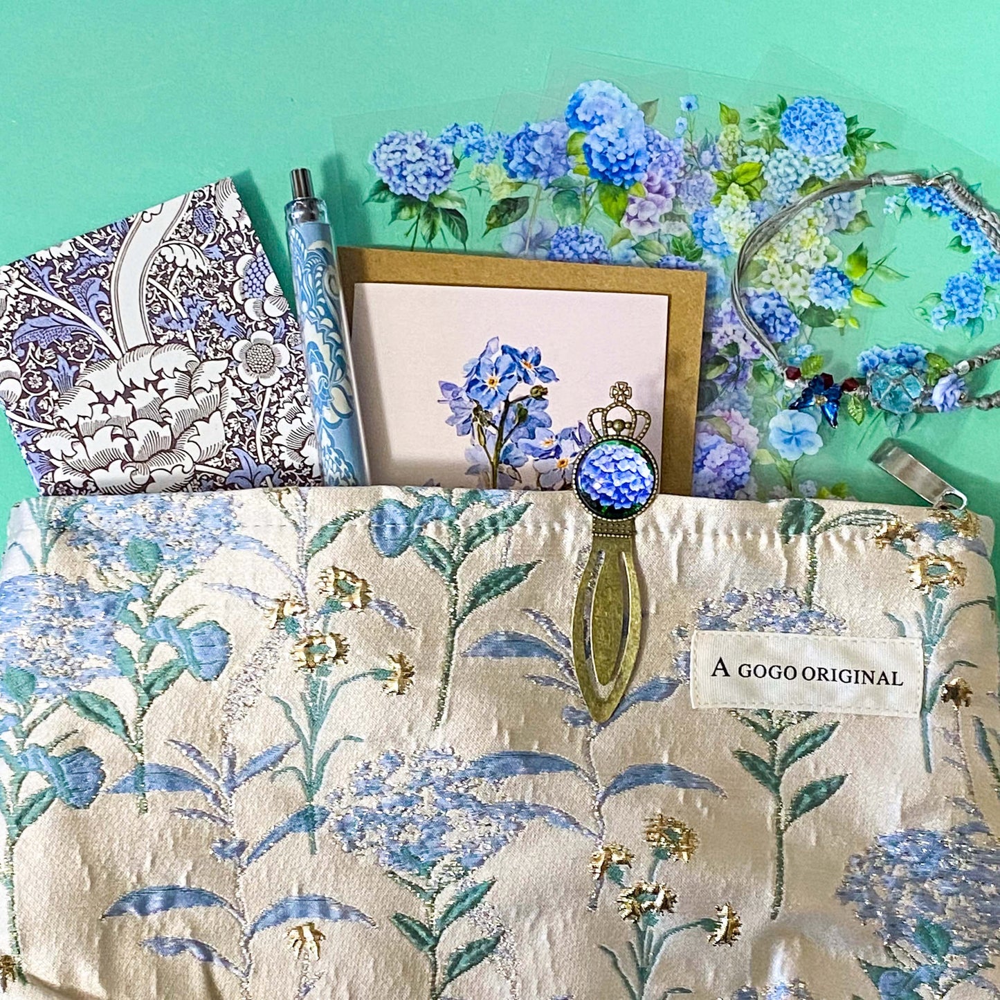 satin hydrangeas letterbox gift set with a lined satin-finish hydrangeas pouch, floral crystal bracelet, notebook, black ballpoint pen, metal bookmark,  2 hydrangea blank note cards and floral sticker sheets. 