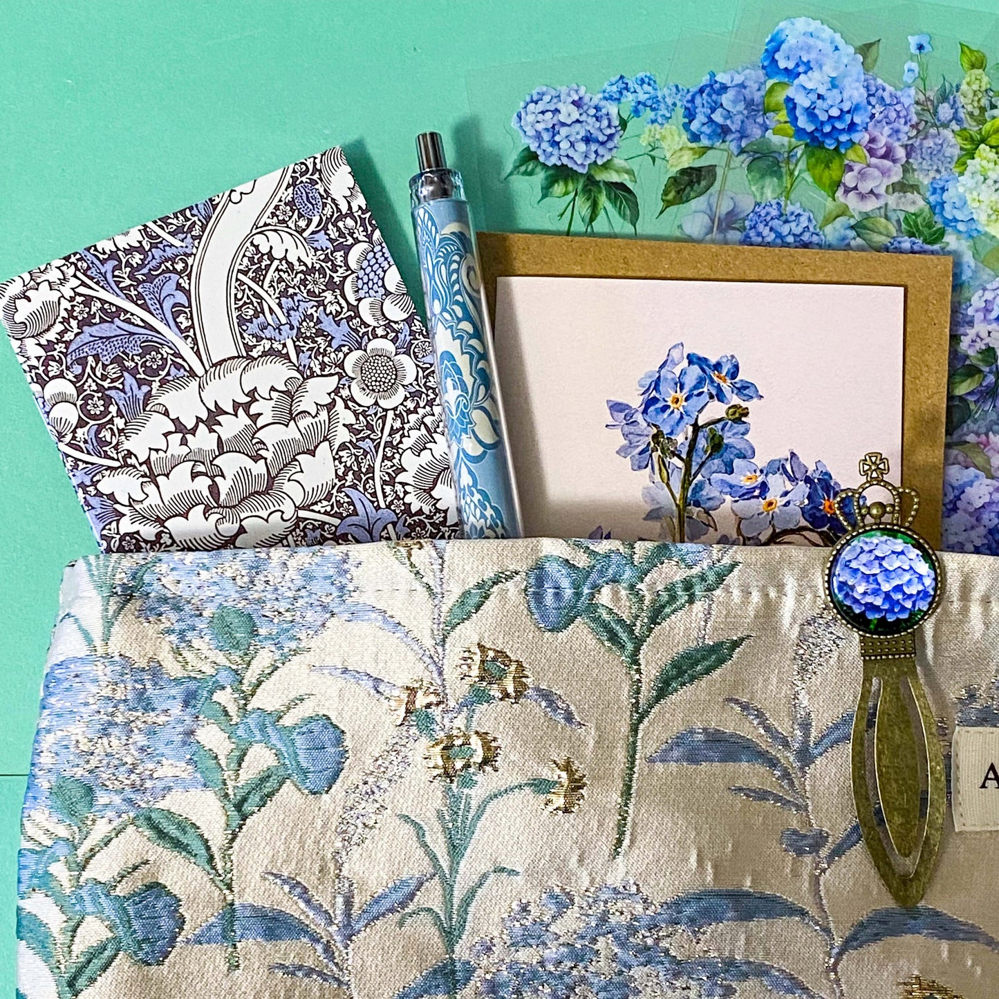satin hydrangeas letterbox gift set with a lined satin-finish hydrangeas pouch, floral crystal bracelet, notebook, black ballpoint pen, metal bookmark,  2 hydrangea blank note cards and floral sticker sheets. 