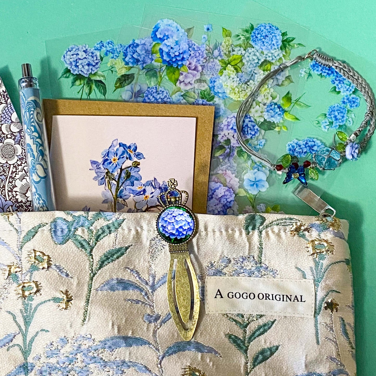 satin hydrangeas letterbox gift set with a lined satin-finish hydrangeas pouch, floral crystal bracelet, notebook, black ballpoint pen, metal bookmark,  2 hydrangea blank note cards and floral sticker sheets. 