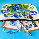 forget me not floral gift set with canvas pouch, botanical note cards, floral pen, bronze owl bookmark and blue crystal bracelet on turquoise background.