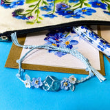forget me not floral gift set with canvas pouch, botanical note cards, floral pen, bronze owl bookmark and blue crystal bracelet on turquoise background.