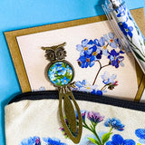 forget me not floral gift set with canvas pouch, botanical note cards, floral pen, bronze owl bookmark and blue crystal bracelet on turquoise background.