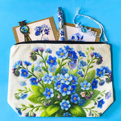 forget me not floral gift set with canvas pouch, botanical note cards, floral pen, bronze owl bookmark and blue crystal bracelet on turquoise background.