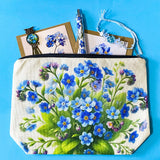 forget me not floral gift set with canvas pouch, botanical note cards, floral pen, bronze owl bookmark and blue crystal bracelet on turquoise background.