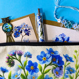 forget me not floral gift set with canvas pouch, botanical note cards, floral pen, bronze owl bookmark and blue crystal bracelet on turquoise background.