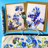 forget me not floral gift set with canvas pouch, botanical note cards, floral pen, bronze owl bookmark and blue crystal bracelet on turquoise background.