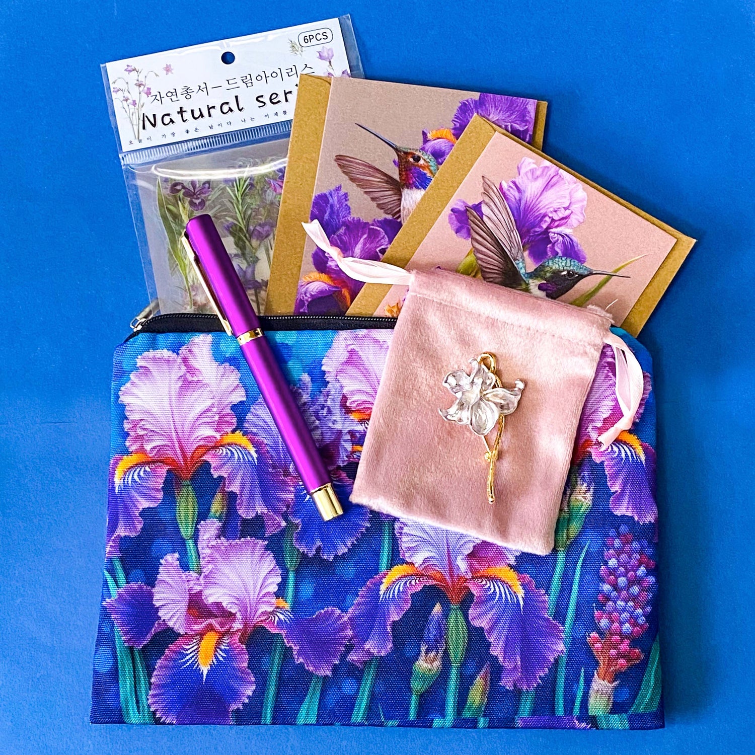 botanical iris letterbox gift set with waterproof floral pouch, purple metal pen, enamel iris brooch in pink velvet bag, clear floral stickers and hummingbird greetings cards. purple, deep jewel blue, lilac and magenta coloured flowers.
