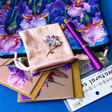 botanical iris letterbox gift set with waterproof floral pouch, purple metal pen, enamel iris brooch in pink velvet bag, clear floral stickers and hummingbird greetings cards. purple, deep jewel blue, lilac and magenta coloured flowers.