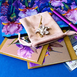 botanical iris letterbox gift set with waterproof floral pouch, purple metal pen, enamel iris brooch in pink velvet bag, clear floral stickers and hummingbird greetings cards. purple, deep jewel blue, lilac and magenta coloured flowers.