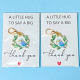 Two packages of keychains with blue bird and wreath design and 'thank you' text on a blue background. The keyring is enamelled on gold tone metal with sparkly rhinestones