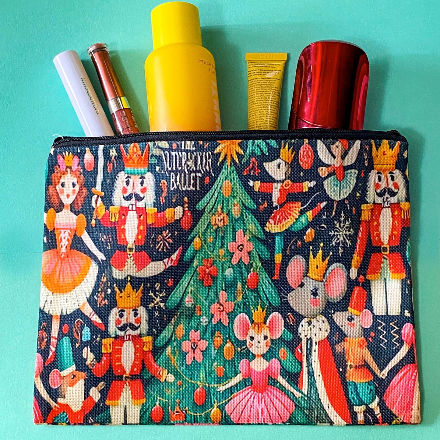A zippered cosmetic pouch with a colorful Nutcracker Ballet theme, featuring illustrated nutcrackers, ballerinas, a Christmas tree, and mice on a dark navy background. Several makeup items are tucked into the top opening against a solid teal background.