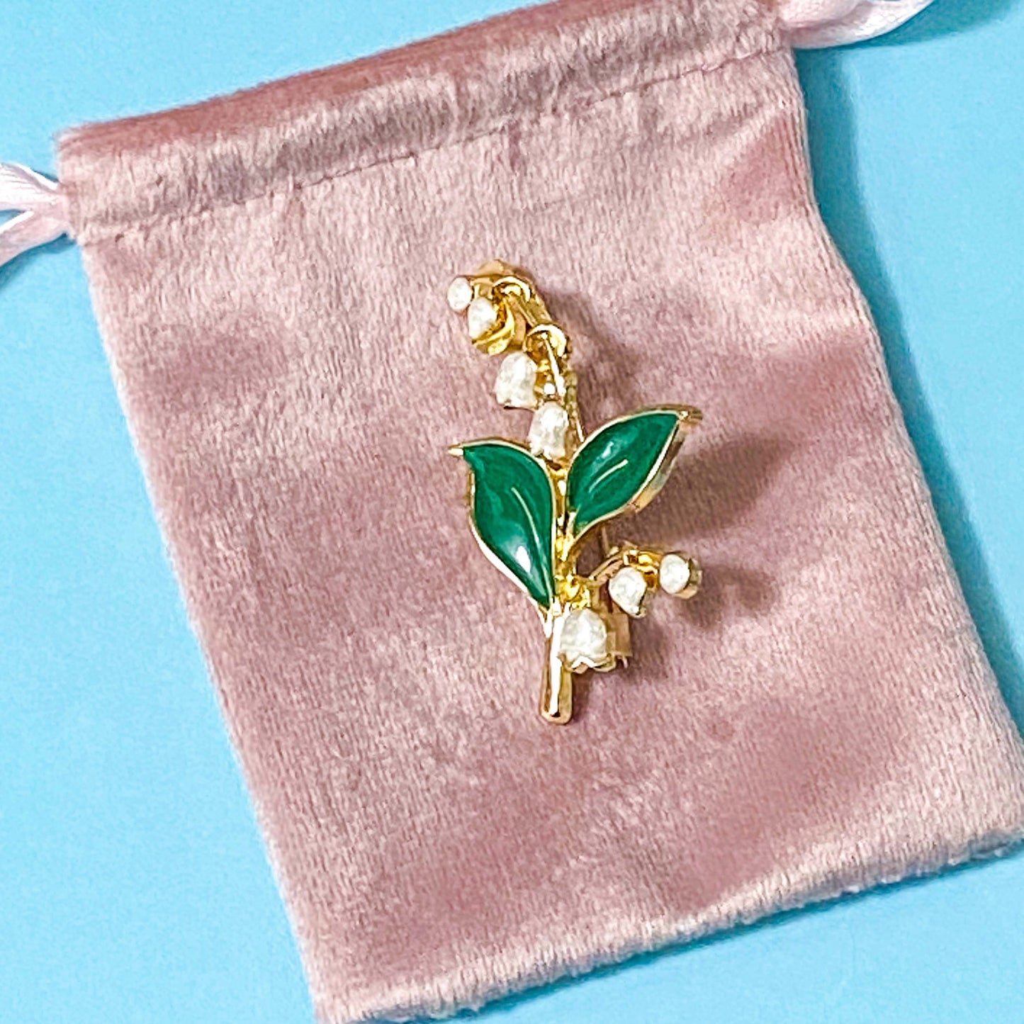 Gold lily of the valley brooch with green leaves and white flowers on a pink velvet bag against a blue background