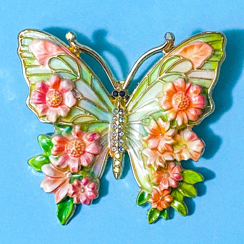 Butterfly-shaped enamel metal brooch with peach and green floral design on a blue background