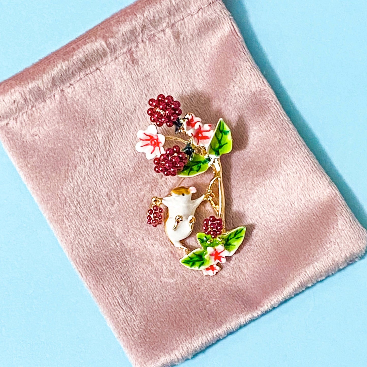 metal and enamel harvest mouse and blackberries brooch on a pink velvet bag on a blue background