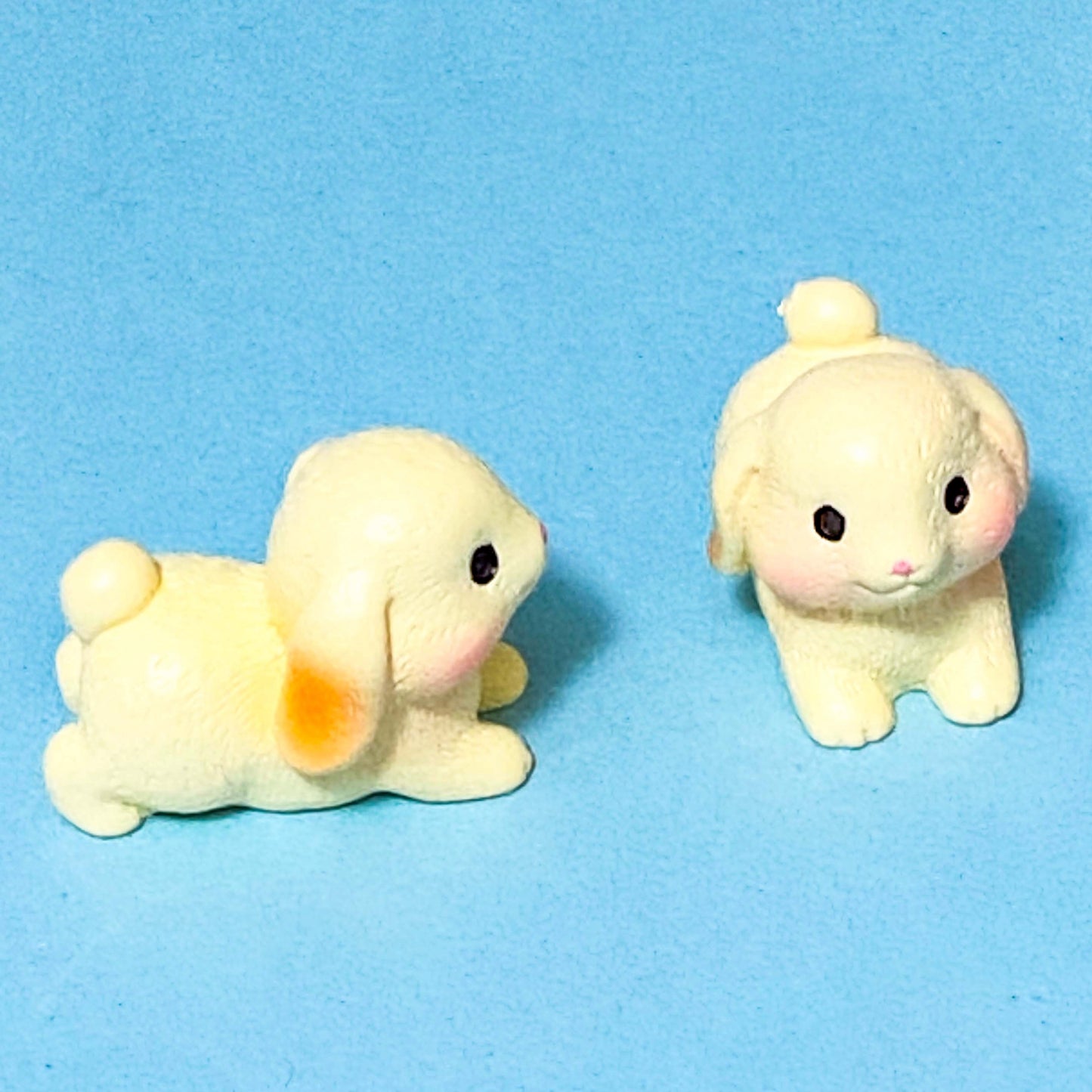 Two small cream 3D miniature animal rabbit figurines on a blue background