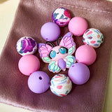pastel silicone bead bundle with butterfly feature bead, pink and lilac round beads and floral patterned beads on a velvet bag