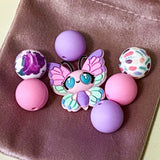 pastel silicone bead bundle with butterfly feature bead, pink and lilac round beads and floral patterned beads on a velvet bag