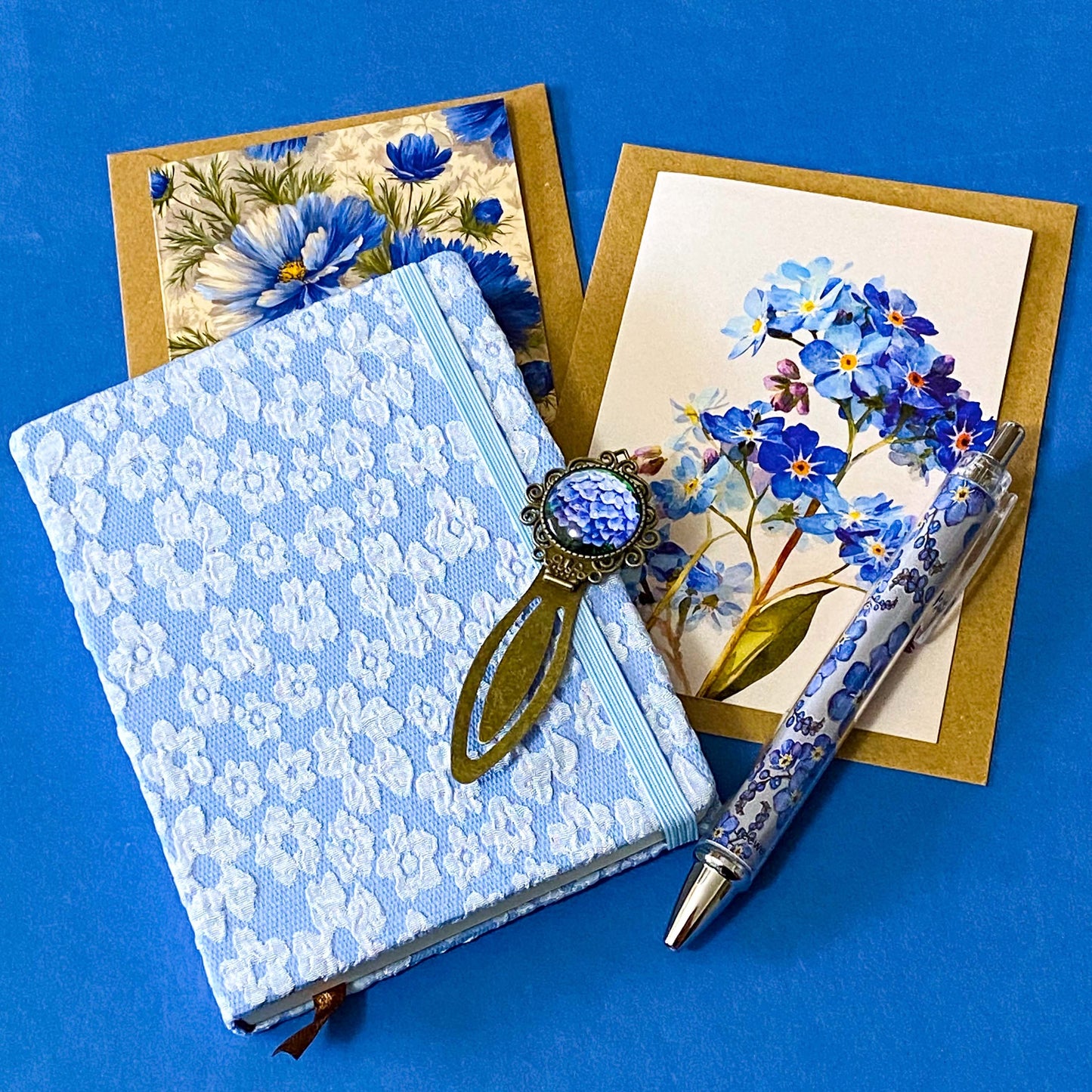 soft blue floral letterbox writing set featuring a textured hardback blue and white floral notebook, matching pen, forget me not cabochon bookmark and blank greetings card and kraft envelope. uk gift shop.