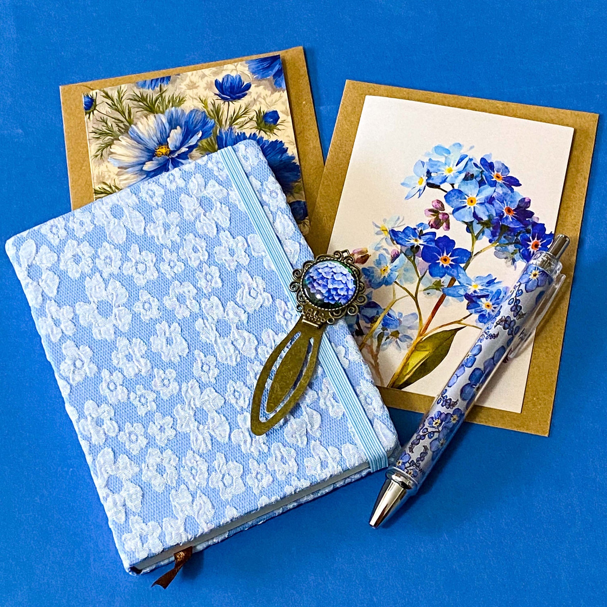 soft blue floral letterbox writing set featuring a textured hardback blue and white floral notebook, matching pen, forget me not cabochon bookmark and blank greetings card and kraft envelope. uk gift shop.